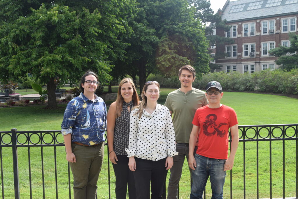 Five people are standing in a row and smiling at the camera. They are in a field on campus, with trees and a brick building in the background. 